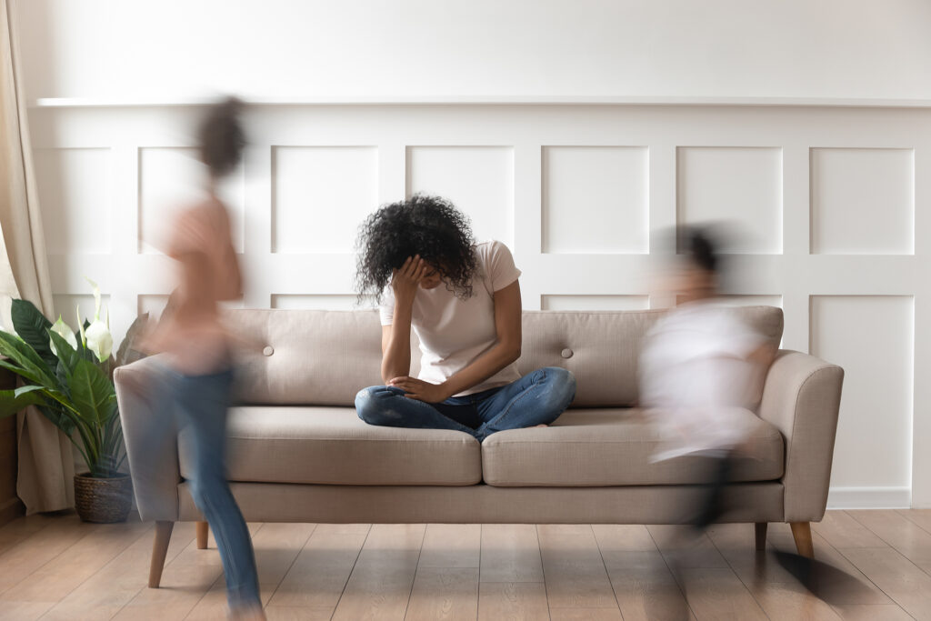 A mother holds her face as her children run around the sofa. This could represent summer parenting stress that therapists in Savannah, GA can offer support with. Learn more by searching for a counselor near me to learn more about Savannah counseling today.
