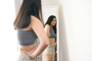 A teenager holds her stomach while looking at herself in the mirror. This could represent summer body issues that therapists in Savannah, GA can offer support with. Learn more by searching for a counselor near me to learn more about Savannah counseling today.