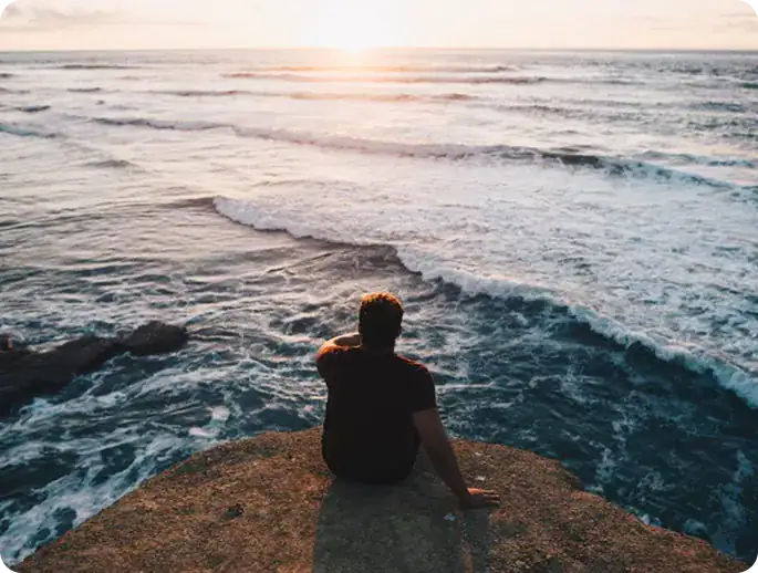 Man sitting on a cliff watching the ocean waves and sunset